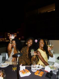 four women sitting at a table with drinks and food in their hands, smiling for the camera