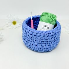 a crochet basket with yarn and knitting needles in it next to daisies