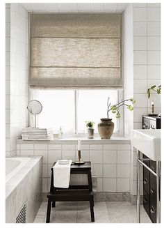 a bathroom with white tile walls and flooring has a window covered in roman shades