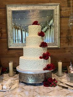 a white wedding cake with red roses on top and candles around it, sitting on a table in front of a mirror