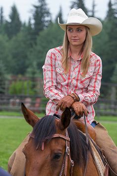 a woman riding on the back of a brown horse in a green field with trees behind her