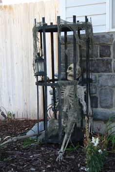 a skeleton in a cage sitting on the ground next to some flowers and plants near a house