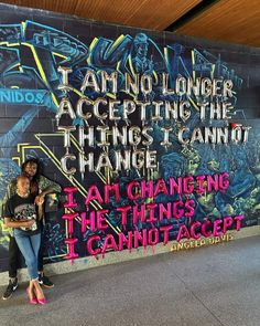 a woman standing in front of a wall with graffiti on it and another person leaning against the wall