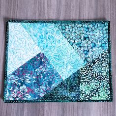 a patchwork placemat with blue and green designs on the top, sitting on a wooden surface