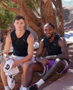 two men sitting on the ground next to each other near a tree and one is holding a water bottle