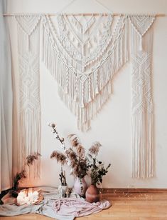 a white wall hanging with tassels and vases filled with flowers on the floor