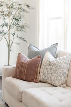 a white couch with several pillows on it in front of a window and a tree