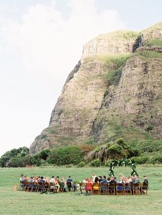 a group of people sitting around a table in the middle of a field with mountains behind them