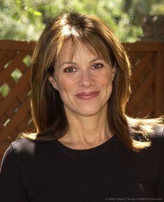 a woman with long brown hair smiling at the camera and wearing a black shirt in front of a wooden fence