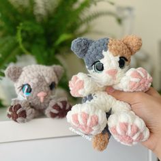 a hand holding two small crocheted kittens in front of a potted plant