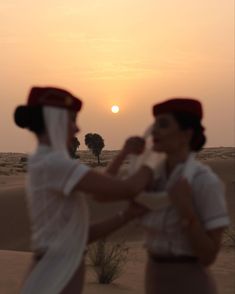 two women dressed in white and red are dancing on sand dunes at sunset with the sun setting behind them