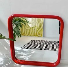 a red mirror sitting on top of a white floor next to a plant in a vase