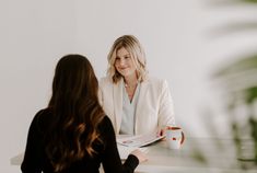 two women sitting at a table talking to each other and one is holding a coffee cup