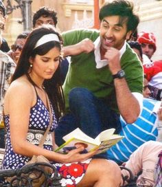 a man sitting next to a woman on top of a bench while holding a book