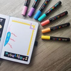 an open notebook with markers and crayons next to it on a wooden table