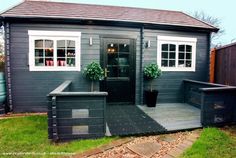 a small gray shed with white windows and plants on the front door, next to a wooden deck