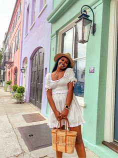 a woman in a white dress and straw hat is holding a wicker basket on the sidewalk