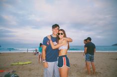 a man and woman standing on top of a sandy beach next to the ocean with people in the background