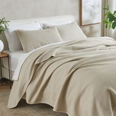 a bed covered in a beige blanket next to a white chair and potted plant