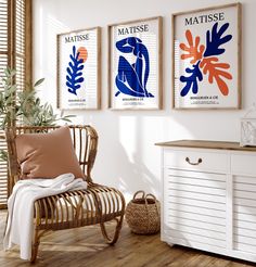 three framed art prints hang on the wall above a wicker chair in a white room