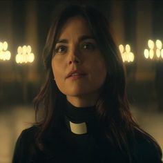 a woman wearing a priest's collar in a dimly lit room with candles behind her