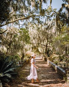 a woman in a white dress and straw hat walking down a path lined with trees