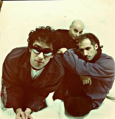 three men sitting next to each other in front of a white wall with one man wearing sunglasses