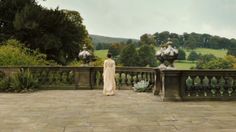 a woman in a long dress is standing on a balcony looking out over the garden