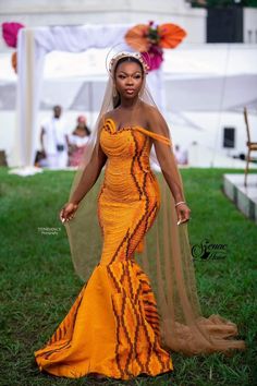 a woman in an orange and yellow dress standing on the grass with a veil over her head