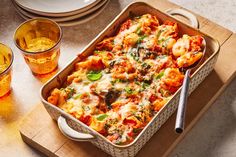 a casserole dish with pasta and vegetables on a cutting board next to drinks