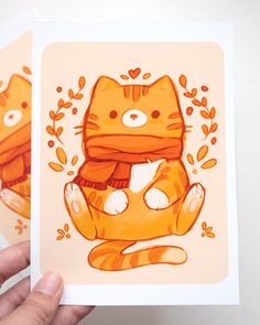 two orange cards with cats on them in front of a white wall and hand holding one