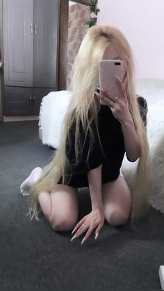 a woman with long blonde hair sitting on the floor looking at her cell phone and holding it up to her face