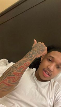 a man with tattoos on his arm laying in bed and pointing at the camera while wearing a white t - shirt