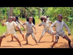 several children are dancing on a dirt road in front of some trees and bushes with their arms stretched out