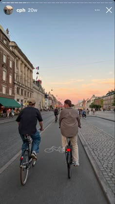 two people riding bikes down the street in front of buildings at sunset or dawn,