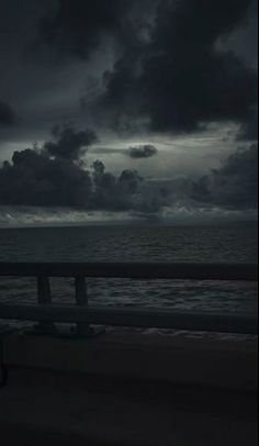 a bench sitting on the side of a beach under a cloudy sky with dark clouds