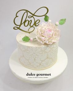 a white cake with a pink flower on top and the word love spelled in gold