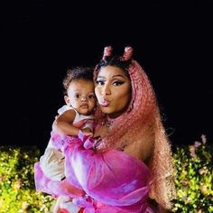 a woman with pink hair holding a baby in front of bushes and trees at night