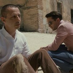 two men sitting on the ground in front of an old building, one is looking at another man