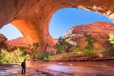 a man standing in the middle of a river under a large rock arch with trees on both sides