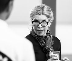 an older woman wearing glasses and holding a glass in her hand while standing next to another person