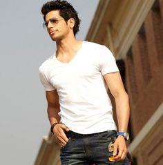 a man in white shirt and jeans standing next to a brick building with his hand on his hip