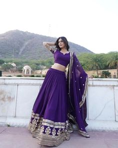 a woman in a purple lehenga poses for the camera with her arms behind her head