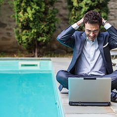 a man sitting on the ground in front of a swimming pool using a laptop computer