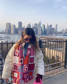 a woman standing on top of a bridge wearing a scarf and mitts with the city in the background