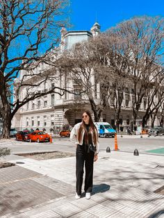 a woman standing on the sidewalk in front of a large building with many trees around her