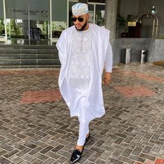 a man walking down the street wearing a white outfit and black shoes with sunglasses on his head