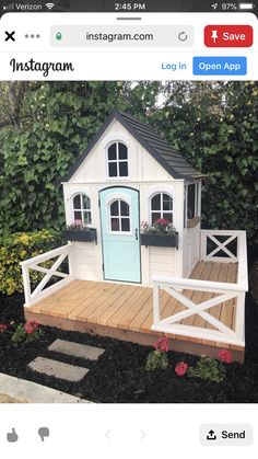 a small white house with a blue door and window on the deck in front of some flowers