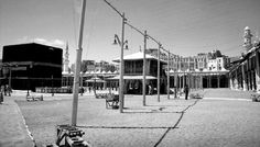 black and white photograph of an empty park with benches in the foreground, buildings in the background