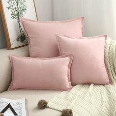 three pink pillows sitting on top of a white couch next to a book and plant